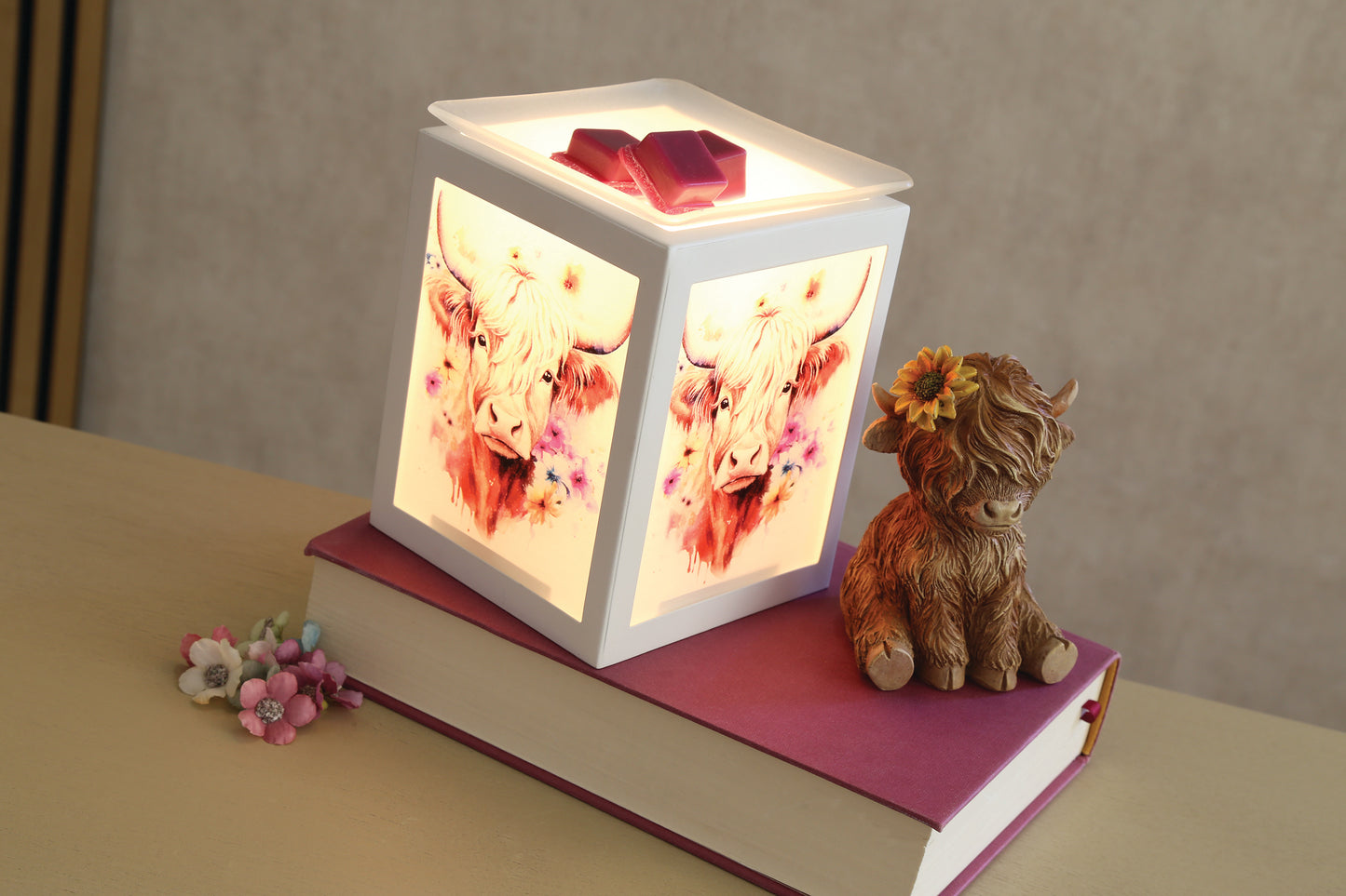 A glowing white cube-shaped wax warmer with painted Highland cow artwork on its sides, displayed on top of a magenta book with a small Highland cow figurine beside it.